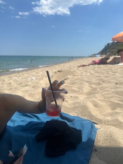A first-person perspective shot from a beach in Byala, Bulgaria, shows a hand holding a clear plastic cup with a red beverage and a black straw. The cup is half full and contains ice and what appears to be a slice of lemon or lime. The hand has light blue nail polish. The person is lying on a blue towel on the sandy beach. A black garment is placed on the towel. In the background, the sea is visible with small waves lapping on the shore. Several people are sunbathing on the beach, some under umbrellas. A green, tree-covered cliff face rises in the distance, characteristic of the Byala coastline. The sky is clear blue with scattered white clouds.