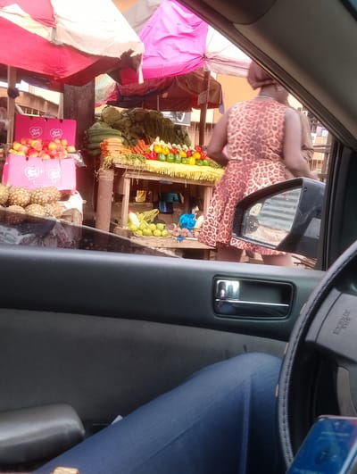 The image captures a street scene in Aketu-Oja, Nigeria, from the perspective of someone inside a vehicle. In the foreground, the interior of the car is visible, including a portion of the steering wheel, a door handle, and the driver's legs clad in blue jeans. Through the car window, a vibrant market stall is in view. Several people are present, with a woman in a leopard-print dress prominently featured near the stall. Various fresh produce, including pineapples, apples, carrots, and peppers, are displayed on wooden tables and in pink crates. The market is covered by colorful tarps, suggesting an outdoor setting. The overall scene depicts a bustling market environment typical of a Nigerian city.