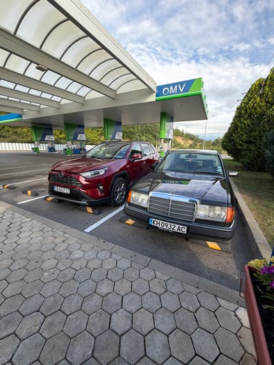 At a gas station identified by an OMV sign in Belo Pole, Bulgaria, two cars are parked. A dark-colored Mercedes-Benz, license plate KH 0932 AC, is positioned in the foreground on the right, angled towards the viewer. To its left and slightly behind is a red Toyota RAV4, license plate CB 8437 CC. Both vehicles are under the canopy of the gas station. The ground in front of the cars is paved with hexagonal concrete tiles, and the area around the cars is marked with white lines and yellow parking blocks. The environment includes lush green trees and a cloudy sky.