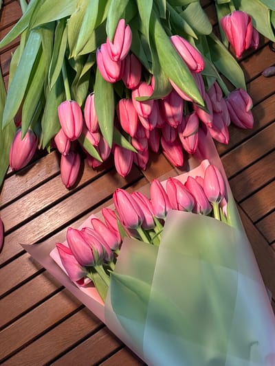 The scene, located in Slavyansk-na-Kubani, Russia, depicts a top-down view of numerous pink tulips arranged on a slatted, reddish-brown wooden surface. No individuals are present.
A large quantity of loose, cut pink tulips with prominent green stems and leaves occupies the upper and left portions of the frame. Their blossoms are primarily closed or partially open, displaying a consistent vibrant pink hue with subtle purple undertones on some petals.
In the lower right section of the image, two distinct bouquets of similar pink tulips are visible. Each bouquet is individually wrapped in translucent paper: one in a light pink or peach-colored material, and the other in a light green or blue-tinted material, obscuring the lower stems.
The wooden surface features parallel slats running diagonally from the top-left to the bottom-right, exhibiting a visible wood grain pattern. The scene is evenly lit, with subtle shadows cast by the tulips.