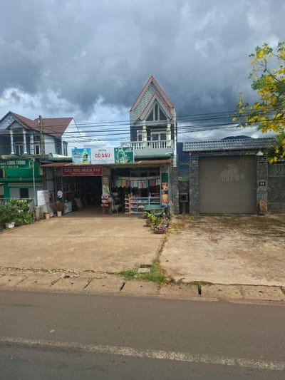 This is a ground-level, eye-level outdoor shot of a commercial street in Tan Phu 2, Vietnam. The sky is overcast with dark, heavy clouds, and the lighting is dim. On the left, a blue building with white trim and columns stands next to a green building with Vietnamese text. In the center, a low-slung establishment features a red sign reading "CẮT TÓC MIỄN PHÍ THỨ 3 HÀNG TUẦN" (Free Haircut Every 3rd Weekday) and displays goods on shelves outside. Further to the right, a large grey metal gate blocks access to a property, with a stone accent and a small sign numbered "04" on the wall. A flowering yellow tree is visible on the far right, with its branches extending over the scene. The ground in front of the buildings is a concrete or asphalt area with scattered debris. The street in the foreground is asphalt with a white line marking.