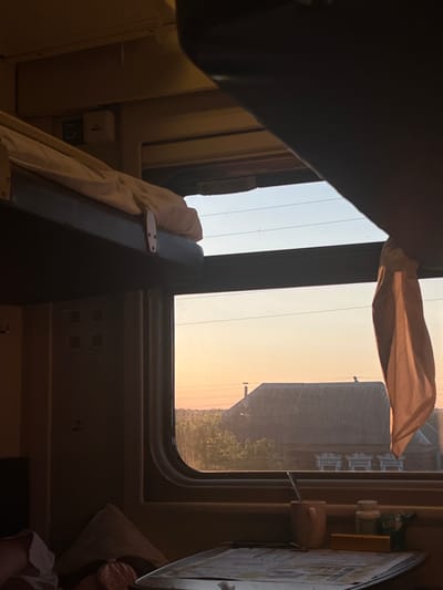 This image depicts the interior of a train compartment at dusk, as indicated by the warm, golden light filtering through the window. The scene is viewed from a low angle, looking up towards the window and the outside landscape.
The main subjects are the window itself, revealing a hazy sky transitioning from light blue to a warm orange, and the silhouette of buildings and trees outside. A light-colored curtain is partially drawn across the top of the window, adding a soft, diffused effect to the light. Inside the train, dark upholstery of seats or bunks dominates the foreground. A white blanket or sheet is draped over a dark surface, secured with a simple metal clasp. Electrical outlets are visible on the wall of the compartment, suggesting a modern train.
The time of day is clearly either sunrise or sunset, given the colors of the sky. The weather appears calm, with no indication of rain or strong winds. The presence of buildings and trees suggests a rural or semi-urban environment outside the train. The overall mood is one of quiet travel and observation, with a sense of transition as day turns to night. There is no visible text in the image. The location context of Анненково-Лесное, Russia, further grounds the scene in a specific geographical setting.