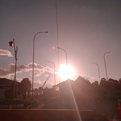 The image captures a street scene in Sihomcot, Indonesia, during sunset. The low sun creates a bright flare in the center of the frame, casting long shadows. In the foreground, a road is visible with traffic cones and a low barrier. Several streetlights stand along the roadside. In the background, buildings and trees are silhouetted against the sky. A tall transmission tower is prominent to the left of the sun. The sky is a gradient of pink and orange, with scattered clouds. No people are visible in the image.
