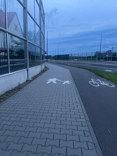 The image depicts a sidewalk and a bicycle lane in the city of Zabrze, Poland, likely during the early morning or late evening due to the dim, overcast sky. The sidewalk is paved with gray interlocking bricks and features a white silhouette of a pedestrian with outstretched arms, indicating it is a designated walking path. Adjacent to the sidewalk is a dark asphalt bicycle lane marked with a white bicycle symbol.
To the left of the sidewalk, a modern building with a facade of floor-to-ceiling glass windows is visible, reflecting the cloudy sky and surrounding structures. A concrete base supports the glass structure. A black metal fence runs along the base of the building.
Beyond the bicycle lane, a road with multiple lanes is present, with white lane markings and a pedestrian crossing indicated by arrows. A distant vehicle is visible on the road. A chain-link fence runs along the side of the road, separating it from the background, which includes trees and more buildings. Streetlights are positioned along the road. The overall environment suggests an urban or semi-urban setting with infrastructure for pedestrians and cyclists.