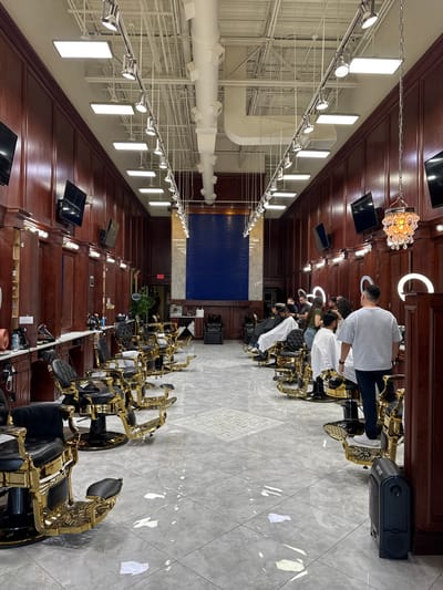 This image captures the interior of a brightly lit, upscale barbershop or hair salon in Carol Stream, United States of America. The scene depicts a long row of barber stations, each featuring an opulent black barber chair with intricate gold-colored frames.
Several individuals are present, with barbers actively providing services to clients. One barber, viewed from behind in a light gray t-shirt, is attending to a client. Other clients are seated in chairs, some draped in white capes, suggesting hair-cutting or styling is in progress.
The shop's design combines classic and modern elements, featuring dark wooden paneled walls and ceiling sections. The floor is covered with modern, light gray tiles. The space is extensively lit by a combination of rectangular LED panels, track lighting with spotlights, and prominent circular LED ring lights at each barber station. A decorative crystal-like chandelier adds to the luxurious ambiance. Exposed white ventilation ducts and structural elements are visible on the high ceiling.
Notable objects include black flat-screen monitors mounted on the wooden panels above each station and a black upright vacuum cleaner branded "eye-vac" on the floor. An "EXIT" sign is visible in the background above a doorway. The overall impression is one of a clean, well-maintained, and stylish establishment where hair care services are performed.