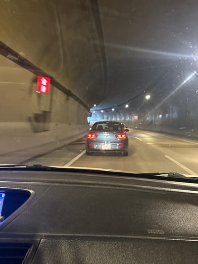 The image shows the interior of a vehicle driving through a tunnel. The view is from the driver's perspective, looking forward through the windshield. A dark-colored sedan is visible in front, with its red taillights illuminated. The tunnel walls are rough and dimly lit by overhead lights. A red traffic signal is mounted on the left wall of the tunnel. The dashboard of the vehicle is partially visible in the foreground. There is no indication of the specific location in Xicotepec, Mexico.