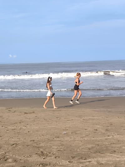 The photograph depicts a beach scene in Tibubeneng, Indonesia. A woman in a white top and skirt walks barefoot along the sandy shoreline, wearing headphones and carrying sandals. Another woman is jogging in athletic attire consisting of a black top and shorts. Both women are positioned in the foreground, with the ocean visible behind them. Waves break along the shoreline, and the sky is a clear, pale blue. An indistinguishable object is visible at sea behind the walking woman. The sand is wet near the water's edge.
