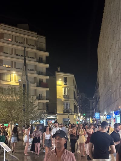 The image depicts a bustling street scene at night in the city of Málaga, Spain, likely in a popular area such as Calle Larios or a nearby side street, given the architectural style and the density of people. The foreground is dominated by a group of people walking and socializing. A man in the immediate foreground wears a pink shirt and a baseball cap. To his right, another man in a black t-shirt is partially visible. In the midground, a crowd of individuals, predominantly women, are dressed in summer attire, suggesting warm weather. Some are engaged in conversation, while others are walking through the street.

The background features multi-story buildings lining the street. On the left, a building with white facades and numerous windows, some illuminated from within, is prominent. A streetlamp is visible in front of this building. Further down the street, a yellow-colored building with ornate balconies and windows is illuminated, creating a warm glow. On the right side of the street, a modern building with a textured, ribbed facade rises vertically, contrasting with the older architecture. Signs for businesses, including an ATM, are visible along the street. The overall atmosphere suggests a lively urban environment at night, with people enjoying the evening.