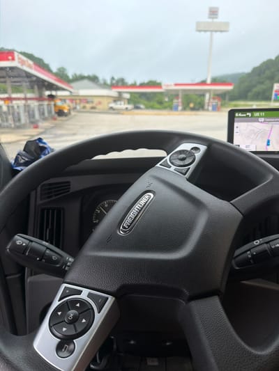 An interior shot from the driver's perspective of a Freightliner truck shows the steering wheel and dashboard. The steering wheel has various controls, including a directional pad and buttons. A GPS device is mounted on the dashboard to the right, displaying a map and traffic information. Outside the truck, a gas station with red and white awnings is visible, along with pumps and a tall pylon sign. Trees and hills form the backdrop under a cloudy sky. The location is identified as Wildwood, United States.