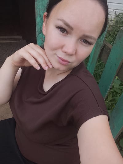 The media file is a selfie of a young woman with dark hair, taken outdoors. She has her hand resting on her chin and is looking at the camera with a slight smile. She is wearing a brown ribbed t-shirt and appears to be in a rural or semi-rural setting, with a wooden fence painted green and lush greenery visible in the background. The lighting suggests it might be daytime. The overall impression is casual and relaxed.