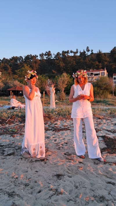 Two women in white outfits and flower crowns stand on a sandy beach at sunset. The woman on the left wears a long white dress with a tiered hem. The woman on the right wears white linen pants and a sleeveless white tunic. In the background, a third woman in a white dress stands near a decorative archway. Other figures are seated on blankets in the distance. The scene appears to be a gathering or celebration on the beach, with a wooded hillside and buildings visible behind the beach. The sky is clear and a pale blue, transitioning to a warmer hue near the horizon, indicating the sun is setting. The sand is littered with dried seaweed and small debris. Torches are visible in the foreground, suggesting the event may continue into the evening.