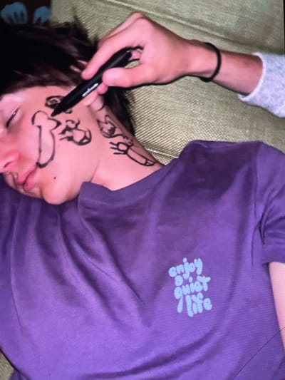 A close-up shot shows a person's face covered in black marker drawings while they lie on a sofa. The drawings include various shapes and figures on the cheek and neck. A hand holding a black marker is seen drawing on the face. The person is wearing a purple t-shirt with a light blue graphic that reads "enjoy a quiet life." The background appears to be a textured green sofa. The image does not contain any information that would place it in Harmanli, Bulgaria.