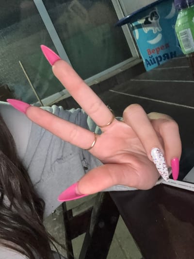 The image shows a close-up of a person's hand making a peace sign. The hand has long, bright pink stiletto-shaped acrylic nails on most fingers, with one finger sporting a white nail with glitter. Two thin gold rings are visible on the fingers. The person is wearing a light grey hooded sweatshirt. The background is blurred but appears to be an outdoor setting, possibly a patio or garden area, with a table and some containers visible. One container has text in Cyrillic script. The image composition suggests the photo was taken by the person themselves. There are no indications of the city of Malo Konare, Bulgaria in the image.