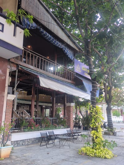 The image depicts a street-level view of an outdoor seating area of a business in Tuy Hòa, Vietnam. The focus is on a multi-tiered structure with a wooden staircase leading to an upper level covered with awnings. A sign with Vietnamese writing is partially visible near the staircase. The structure appears to be built around a large tree. The seating area includes black metal chairs and tables arranged along a stone pavement, with plants and flowers providing decoration. The surrounding buildings are visible in the background, along with some street traffic.