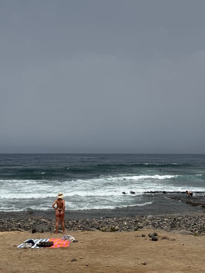 This media file depicts a beach scene in Arona, Spain. The image is oriented vertically. On the right side of the frame, a sandy beach transitions to a rocky shoreline, where the ocean meets the land with breaking waves. In the foreground, a person wearing a bikini and a straw hat stands on the sand, facing towards the ocean. Next to them, a colorful striped towel is laid out on the sand, with some clothing and a bag placed on it.
The ocean is active with white-capped waves rolling towards the shore. Several surfers can be seen in the water further out. The sky is overcast, indicating a cloudy or possibly stormy day, casting a muted light over the scene. The overall impression is one of a somewhat subdued beach day, with the focus on the solitary figure on the sand and the dramatic ocean.