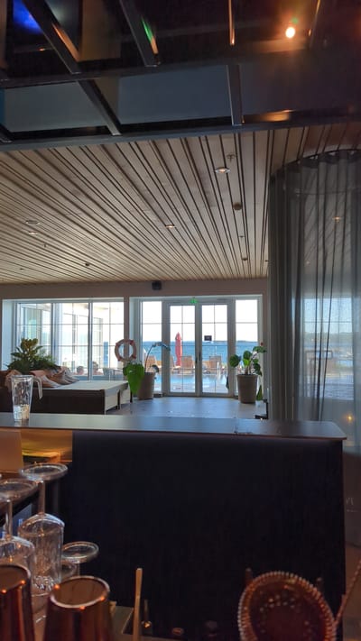 The image depicts a bar area looking out towards a coastal view in Larkollen, Norway. In the foreground, a dark bar counter is visible with several cocktail glasses and bar tools. Beyond the counter, a large glass-paneled wall offers a view of an outdoor seating area with lounge chairs, a red umbrella, and a swimming pool, leading to the sea. Two potted plants are placed near the glass doors. Inside, a seating area with cushions and a life preserver is arranged. The ceiling features a wooden slatted design with recessed lighting. The overall ambiance suggests a contemporary resort or hotel bar.