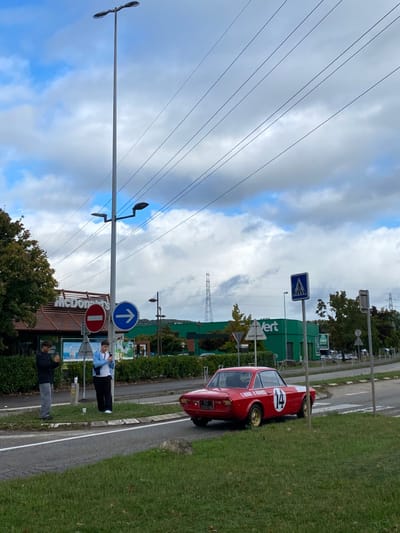 The image captures an outdoor daytime scene in Dorlisheim, France, under a partially cloudy sky. A red vintage Lancia Fulvia Coupé, displaying a white circular racing number "14" on its side, is positioned on a paved road facing away from the camera. Its rear license plate, partially visible, reads "CZ-285-HA," and it features gold-colored wheels.

To the immediate left of the car, on a paved median or sidewalk, two males are standing. The male on the left, wearing a dark long-sleeved top and grey pants, appears to be observing something in his hands. The male on the right, dressed in a light blue jacket over a white top and dark pants, is actively engaged with a smartphone; a white disposable cup rests on the ground near his feet.

In the background, a McDonald's restaurant with its distinctive red-brown roof and signage is visible to the left. Further to the right, a large green building with prominent "VERT" signage stands. A tall, multi-lamp street light pole dominates the central frame, from which several power lines extend horizontally across the sky. Multiple road signs are present, including a red circular 'No Entry' or 'Do Not Enter' sign, a blue circular 'Mandatory Right Turn' arrow, and a blue square 'Pedestrian Crossing' sign. In the far distance, a large metal electricity pylon is visible against a backdrop of distant hills. The foreground consists of a grass verge with scattered rocks, transitioning to a paved road surface with clear lane markings. No direct interactions are observed between the individuals and the vehicle.