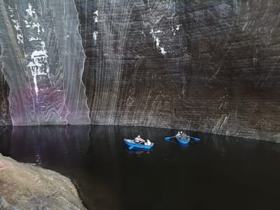 The image depicts two small blue rowboats with people inside navigating a dark, still body of water within a cavernous space. The cavern walls are rough and textured, with varying shades of gray and white, suggesting a salt mine. In the foreground on the left, a textured, light-colored material, possibly netting or fabric, lines a portion of the bank. The water reflects the cavern's textures and the boats.
In the closer boat, a man wearing a grey shirt and a woman wearing a dark top are seated, with the man appearing to be rowing. In the second boat, further in the distance, two individuals are visible, one rowing with blue oars. The overall atmosphere is serene and somewhat mysterious due to the dark environment and the quiet activity. The lighting is dim, typical of an underground location. The scene suggests a tourist attraction within the Turda Salt Mine in Romania, where visitors can experience the unique environment by boat. There is no visible text in the image.