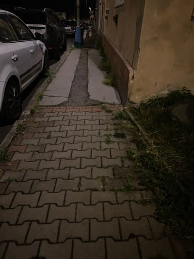 The image shows a sidewalk made of interlocking concrete pavers in Bílina, Czechia. To the left of the sidewalk, there are two cars parked along the street. The first car, closer to the foreground, is white. The second car is darker in color. The sidewalk is bordered on the right by a building with a light brown, weathered facade, and a red brick base. Overgrown grass and weeds are visible along the edge of the sidewalk and between the building and the sidewalk. A blue trash receptacle is visible further down the sidewalk. The scene appears to be from nighttime or early evening, indicated by the artificial lighting and shadows.