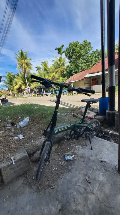 A dark green Pacific folding bicycle is parked on a concrete surface next to a dirt area with sparse dry grass and scattered debris. The bicycle is upright, supported by its kickstand. In the background, there is a road with a few buildings and palm trees. The sky is blue with scattered white clouds. The scene appears to be outdoors in a tropical or subtropical location.