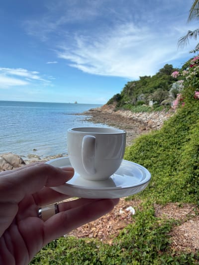 A hand wearing a ring holds a white coffee cup and saucer in the foreground. Behind the cup, a body of water, likely the ocean, is visible. A rocky shoreline with green foliage and pink flowers lines the water. A lush green hillside with trees and palm fronds rises in the background. The sky is blue with scattered white clouds. This scene is located in Choeng Mon, Thailand.