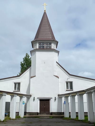 The image shows the exterior of a white church with a brown, conical steeple topped by a gold cross. The church has a prominent entrance with double brown doors. There are two windows on either side of the main entrance, under the eaves of the building. A white colonnade with a slanted roof runs along the front of the building. The sky is cloudy and overcast. The church is located in Sortavala, Russia.