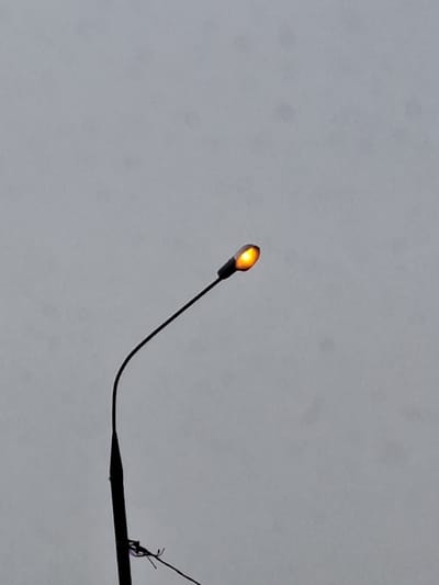 A single street light, located in Obukhovo, Russia, is depicted against an overcast, light grey sky. The dark, slender pole of the street light extends upwards from the lower left portion of the frame, supporting a dark, curved arm that culminates in an elongated, tear-drop shaped lamp housing. The lamp is actively illuminated, emitting an orange-yellow light. Near the base of the pole, several dark wires, some exhibiting faint purple hues, are visible and appear to be secured or tangled. No other structures, individuals, or environmental elements are discernible within the frame.
