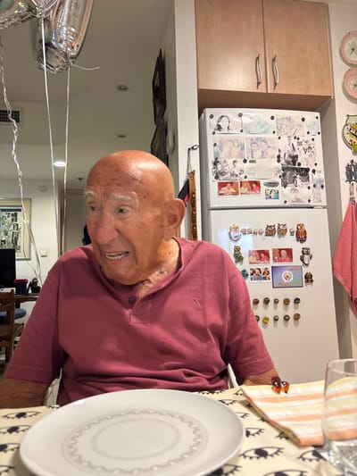 An elderly bald male, appearing to be of advanced age, is seated at a table in a domestic setting, identified as a residence in Bnei Dror, Israel. He wears a short-sleeved maroon Henley-style shirt and is positioned facing slightly to his left, with his mouth open, indicating speech or a reactive expression.

In the immediate foreground, a white circular plate with a decorative rim pattern rests on a tablecloth featuring a repetitive dark eye-like motif. To the right of the plate, a folded orange-and-white striped napkin holds a small amber-colored translucent object, and a clear drinking glass is positioned next to it.

The background on the left displays several silver helium balloons with white ribbons suspended near the top of the frame. Further back, a room is visible with a dark-colored computer monitor on a desk and multiple framed photographs on a wall. The background on the right is dominated by a white two-door refrigerator heavily adorned with numerous photographs and various magnets, including a collection of owl-shaped magnets. Above the refrigerator are two light-colored wooden kitchen cabinets with silver handles. To the right of the refrigerator, a pink towel hangs on a hook, and two decorative, light-colored plates with floral designs are mounted on the wall. Recessed lighting fixtures are visible on the ceiling.