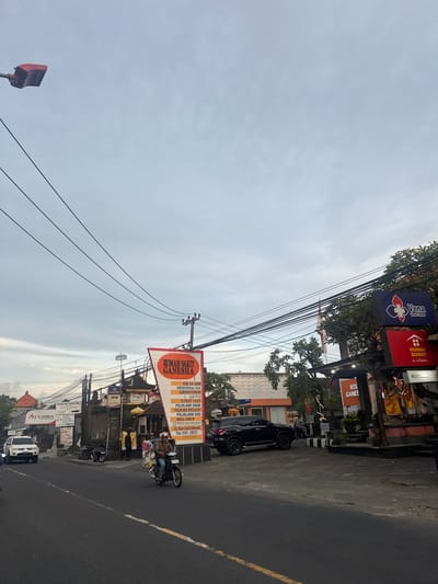 The image shows a street scene in Celuk, Indonesia. A road stretches through the foreground, marked with a yellow dividing line. On the right side of the road, a black car is parked. A motorcycle with two people onboard is positioned nearby, with items piled on the motorcycle. Banners and signs are displayed along the roadside, including "RUMAH SAKIT GANESHA," which translates to Ganesha Hospital, listing services. Other signs include "Ary's mikro" and "BANK BRI ATM." Power lines stretch across the sky. The sky is overcast. The businesses on the street include RSU GANESHA and Vena Wasir Center.