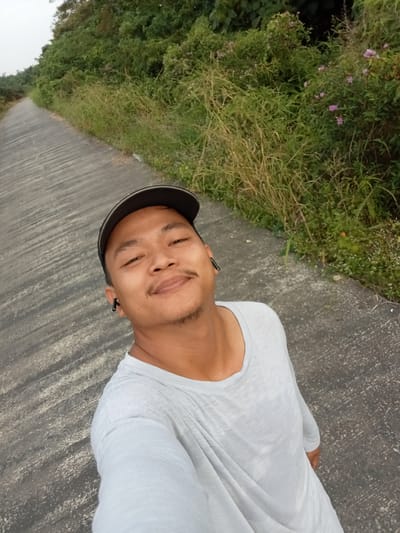 A man wearing a gray long-sleeved shirt and a dark baseball cap is taking a selfie outdoors. He has a mustache and a soul patch. He is wearing black wireless earbuds. He is standing on a paved pathway that curves to the left and is bordered by lush green vegetation and some small purple flowers. The pathway appears to be in a rural or semi-rural area, possibly on the outskirts of Kualu, Indonesia. The sky is overcast.