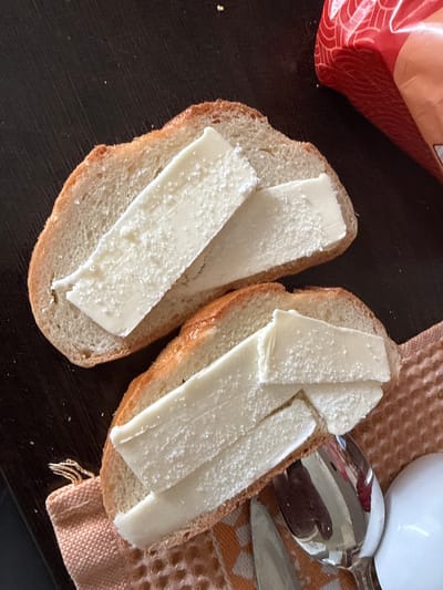 The image shows two slices of bread with slices of white cheese on top, resting on a dark, textured surface. A textured, peach-colored cloth is visible to the right of the bread, partially underneath it. A spoon with a reflective surface, likely holding some liquid, is on the cloth, and a small part of a white mug or bowl is also visible. To the upper left of the bread, a portion of a red and orange bag with a patterned design is visible. The lighting suggests it may be daytime, and the overall scene appears to be a simple meal or snack preparation. There is no visible text in the image.