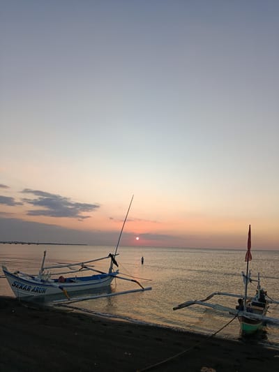 The image depicts a serene sunset scene at a beach in Jangkar, Indonesia. Two traditional Indonesian fishing boats are visible, one closer to the shore and the other further out in the water. The boat on the left, named "SEKAR ARUM" as indicated by blue text on its hull, is white with blue trim and has a wooden outrigger. The second boat, on the right, is smaller and partially submerged, with a green and brown hull and a red flag at its mast.
The sun is setting on the horizon, casting a warm, orange glow across the sky and water. The sky transitions from a pale blue at the top to soft pinks and oranges near the horizon. A few wispy clouds are scattered in the sky. The sea is calm, reflecting the colors of the sunset. In the distance, a single person stands in the shallow water, silhouetted against the setting sun. Further back, a long pier or breakwater stretches across the water, with a line of small structures along it.
The beach itself is dark and sandy, with a gentle wave lapping at the shore. The overall mood of the image is peaceful and tranquil, capturing the quiet beauty of a coastal sunset. The time of day is clearly evening, and the weather appears to be clear and calm. The presence of the fishing boats and the pier suggests a coastal fishing community.