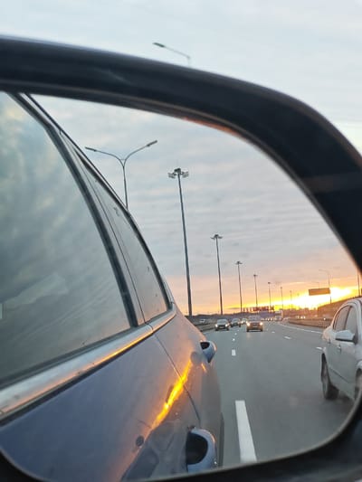 The image, taken from inside a vehicle via the side-view mirror, depicts a highway scene at sunset, potentially located near or in Poroshkino, Russia, based on the presence of infrastructure similar to those in the area. The highway is filled with traffic, indicated by multiple cars visible in the mirror's reflection. Street lights line the highway. The sky is characterized by the colors of a sunset. The left side of the frame is dominated by the side of the vehicle, and a white car occupies a portion of the right side. The presence of other cars and highway infrastructure suggest it is a busy area, potentially a commuter route near Poroshkino.