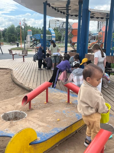 The image depicts a group of children and an adult in a playground area, likely located in the city of Oktyabrsky, Russia. In the foreground, a young child, appearing to be a toddler, is dressed in a tan sweatshirt and tan ribbed pants, with a yellow bucket in their right hand. They are standing on a sand-covered surface with blue and yellow playground structures. Several other children are gathered around a wooden deck in the background, engaged in various activities. Some appear to be looking down at papers or objects. The playground features red pipes, a circular opening in the sand, and wooden benches. In the middle ground, there are larger playground structures with orange and blue geometric shapes and slides. The background reveals residential buildings and trees under a cloudy sky. The overall scene suggests a casual, outdoor gathering or activity in a public space.