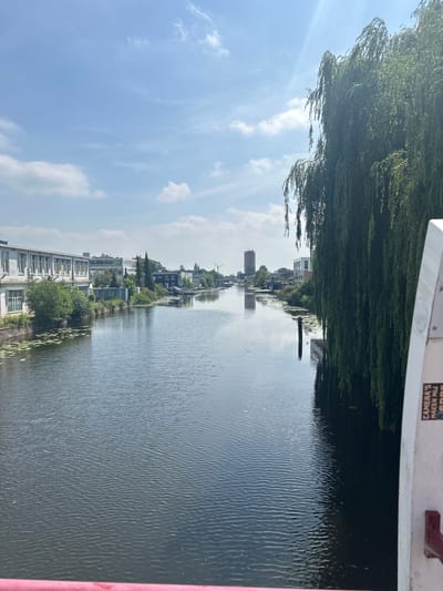 The image depicts a canal in Leiden, Netherlands, viewed from a bridge. The canal runs through an urban or semi-urban area with buildings lining its left bank. These buildings appear to be a mix of industrial or commercial structures, with a long, white building with many windows being prominent on the left. On the right bank, a large weeping willow tree dominates the foreground, its branches cascading down towards the water. The canal itself is calm, with ripples on its surface reflecting the sky and surrounding scenery. The sky is blue with scattered white clouds. In the distance, more buildings and trees are visible, along with what appears to be a wind turbine, suggesting an area that may blend industrial and residential or recreational spaces. The overall scene suggests a typical canal view in a Dutch city.