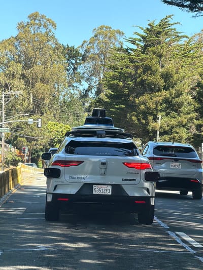 This image captures a sunny day in South San Francisco, California, United States of America. The foreground is dominated by the rear of two light-colored autonomous vehicles, specifically Jaguar I-PACE models, driving on a paved road. The lead vehicle, closer to the camera, displays "WAYMO" on its roof-mounted sensor array and "Ride today" and "TCP 38152A" on its rear. Its license plate reads "53512F3". The second vehicle behind it has a license plate "9GXC945". Both vehicles have their brake lights illuminated. The road has painted lane markings and visible tire marks.
To the left of the road, there is a wooden retaining wall and a yellow guardrail. Beyond that, a street sign indicates "Coronado Drive". Traffic lights are visible in the distance, with one appearing red. A scattering of small orange objects is present on the road surface near the retaining wall.
The background consists of lush green trees with sunlight filtering through their leaves, suggesting a warm and clear day. The sky is a vibrant, cloudless blue. Overhead, power lines are visible against the trees. The overall scene depicts autonomous vehicles operating in a residential or suburban environment characterized by natural landscaping and road infrastructure.