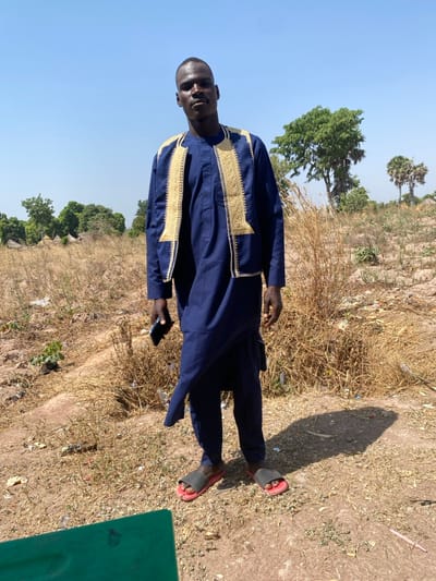 A dark-skinned male, approximately in his twenties to thirties, stands facing forward, looking directly at the camera. He wears a dark blue traditional garment consisting of a tunic and trousers, with a gold-embroidered vest worn over the tunic. He holds a dark-colored smartphone in his left hand, down by his side. His feet are clad in black and red slide sandals.
The individual is positioned outdoors on dry, light brown soil with sparse, straw-colored vegetation and scattered debris, including what appears to be plastic. In the background to the left, several small, round, earth-colored structures with conical roofs are visible amidst trees, suggesting a rural settlement. To the right, mature green trees and two distinct palm trees stand against a clear, light blue sky. Strong overhead sunlight creates a distinct shadow of the individual on the ground to his right. A partial green object is visible in the lower left foreground. The scene is located in Zaki Biyem, Nigeria.