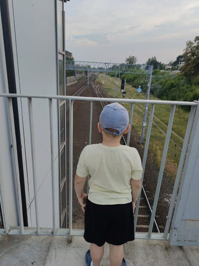 A young boy, facing away from the camera, stands on a pedestrian bridge overlooking a set of train tracks. He is wearing a light yellow t-shirt, black shorts, and a light blue baseball cap. The bridge has a light gray metal railing. In the distance, the train tracks curve to the left, with a railway signal displaying a red light. Overhead, a metal structure spans the tracks, likely part of the railway infrastructure. The scene is set during the day, with an overcast sky. Green trees and vegetation line the sides of the tracks. The boy appears to be observing the train tracks, possibly waiting for a train. The location is identified as Nowy Dwór Mazowiecki, Poland.