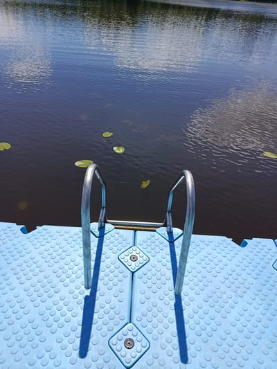 A floating dock made of interlocking blue plastic modules, featuring a textured surface with circular bumps, is positioned at the edge of a dark body of water. A metal ladder with silver handrails extends from the dock into the water. Several lily pads are visible on the surface of the water. The sky above is reflected in the water, showing clouds and blue sky. The scene is outdoors, likely in a natural setting, consistent with the location in Мяжанскі сельскі Савет, Belarus. There are no people visible in the image.