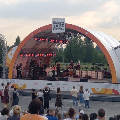 The image shows an outdoor concert stage set up for the Moscow Jazz Festival. A band is performing on stage, consisting of a female singer, a male guitarist, a keyboardist, a drummer, and another guitarist. The stage is adorned with red lights and a large banner at the top reads "Moscow Jazz Festival." In front of the stage, a crowd of people are gathered to watch the performance. The background shows trees and a cloudy sky. The location of the event is stated as Путрышкаўскі сельскі Савет, Belarus.