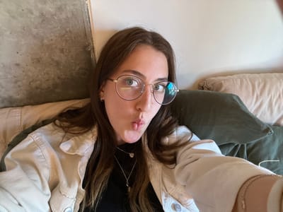 A person with light skin, wearing round glasses, a black top, a light beige jacket, and a silver necklace, is positioned upside down in the frame. They are making a kissing face. Two pillows, one light beige and one olive green, are partially visible on the left. The background consists of a white surface and a gray concrete floor.