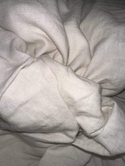 The image displays a close-up, top-down perspective of extensively folded and creased light-colored textile, specifically appearing off-white or cream. The fabric exhibits a woven texture with visible individual threads, consistent with a natural fiber such as cotton or linen. Numerous soft undulations, deep folds, and irregular wrinkles create varying patterns of light and shadow across the entire frame. No distinct patterns, foreign objects, individuals, or environmental elements are visible other than the fabric itself. The scene is located in the city of Wealden, United Kingdom.