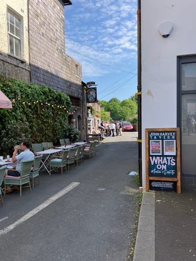 The scene is a street-level view of John Harvey Tavern in Lewes, United Kingdom. The view is framed by an archway. An outdoor seating area with green chairs and white tables extends along the right side of the street. Some people are seated at the tables. A sign advertising "John Harvey Tavern" and "What's On Music & Events" is affixed to the building on the right side of the street. Further down the street, a group of people stand or sit outside the pub. The sky is partially visible with scattered clouds. A sign with "HARVEYS LEWES" is visible above the pub.