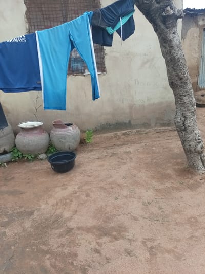 In Zinna, Nigeria, a domestic scene unfolds outside a simple dwelling. Clothes, including a blue t-shirt with white lettering that reads "AIRWAYS" and a pair of blue athletic pants with white stripes, hang drying on a clothesline. Nearby, two large, round ceramic pots, one with a white rim and the other partially broken, stand on the sandy ground. A black plastic basin rests in front of the pots. The wall behind the drying clothes is adorned with graffiti. A large tree trunk with rough, textured bark stands prominently to the right of the frame, its branches extending upwards. The ground is composed of reddish-brown sand, with sparse patches of dry grass. The overall impression is one of everyday life in a rural or semi-urban Nigerian setting.
