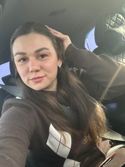 The scene is located within a vehicle in the city of Mostovaya, Russia. A female individual with long, dark brown hair parted down the middle is seated, occupying the foreground of the image. She has a fair complexion, distinct dark eyebrows, brown eyes, and light pink lipstick on her lips. Her right hand is raised, with fingers splayed, resting lightly on the top of her head in a posed manner. She is wearing a dark brown long-sleeved cardigan featuring a white and maroon argyle pattern on the chest. A dark seatbelt is secured across her torso. On her right ear, a small, gold-colored stud earring with an intricate design is visible. In the lower right of the image, a brown or metallic lipstick pen is partially visible. The interior of the vehicle shows a light-colored ceiling and a dark, quilted headrest in the background to the right. Sunlight enters from the right side of the frame, illuminating part of her hair and sleeve. A blurry bright blue sky is visible through the window on the right.