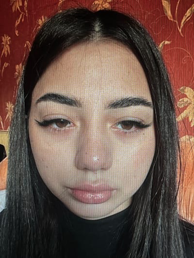 The image shows a close-up, head-on shot of a young woman with dark hair, light brown eyes, and full lips. She is wearing a black garment. The background is a red, patterned wallpaper. The image does not provide any information about the city of Chotěšov, Czechia, or any specific location.