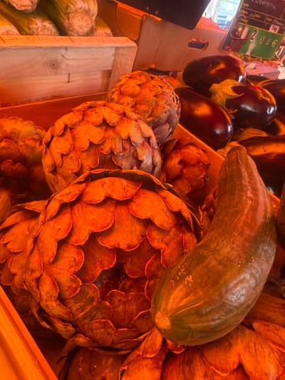 In Saint-Claude, France, at a market stall, a close-up shot captures a variety of fresh produce. Dominating the foreground are several large artichokes, their outer leaves a warm, earthy orange hue, revealing intricate layers. To the right, a long, dark green zucchini lies diagonally across the artichokes. In the background, more artichokes are visible, alongside glossy, dark purple eggplants. A wooden crate with a visible label and some loose celery stalks add to the market scene. The lighting casts a warm, golden glow over the entire arrangement.