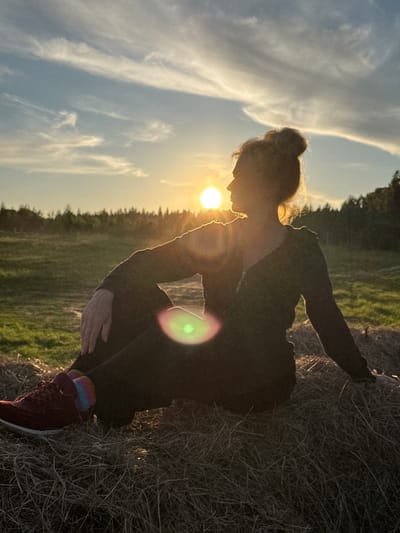 This image captures a serene outdoor scene at sunset in Стеклянный, Russia. The low, golden sun is prominently featured in the upper left, casting the main subject into a strong silhouette and creating a warm, reflective atmosphere.

The central subject is a person, likely female, in a crouched or squatting position, facing slightly away from the camera. Her hair is styled in a bun, clearly visible in silhouette against the bright sky. She is dressed in dark, long-sleeved attire, with her hands visible resting on her legs or the ground. A notable detail is the colorful, rainbow-striped socks she wears, offering a vibrant contrast to her dark shoes and clothing. She appears to be leaning against or observing a mound of dry grass or hay, which occupies the right side of the frame.

The background features a vast, clear sky adorned with wispy, elongated clouds, particularly noticeable on the left. A horizon line of dark trees or a forest can be seen in the distance, indicating a rural or natural landscape. The sun's intense light generates a distinct lens flare, manifesting as a green and magenta circular glow to the right of the person's silhouette. The overall mood is peaceful and contemplative, highlighting the beauty of the golden hour. No text is visible within the image.