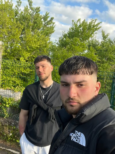 Two young men are posing outdoors. The first man is in the foreground, wearing a black "The North Face" vest. The second man is behind him wearing a black T-shirt and a silver cross necklace. The background is a green chain link fence and trees. The location is the city of Knockaunbaun, Ireland. The sky is blue with white clouds.