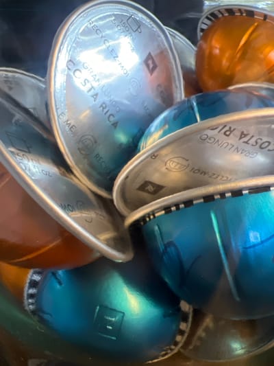 This image is a close-up, slightly blurry, overhead view of a pile of Nespresso coffee capsules. The capsules are of various colors, including orange, blue, and green, and are layered on top of each other in a container. Some capsules have visible text printed on them, including "GRAN LUNGO," "COSTA RICA," "RECYCLEZ-MOI," and "150ml 15.07 FL.OZ." There are also Nespresso logos visible on some of the capsules. The lighting suggests an indoor setting, and the focus is on the texture and colors of the capsules. There are no people or discernible environmental clues to suggest a specific time of day, weather, or emotions. The location context provided is Opglabbeek, Belgium.